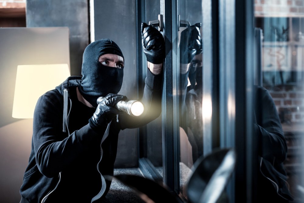 A masked burglar at night inspecting ways to enter through a window while holding a flashlight